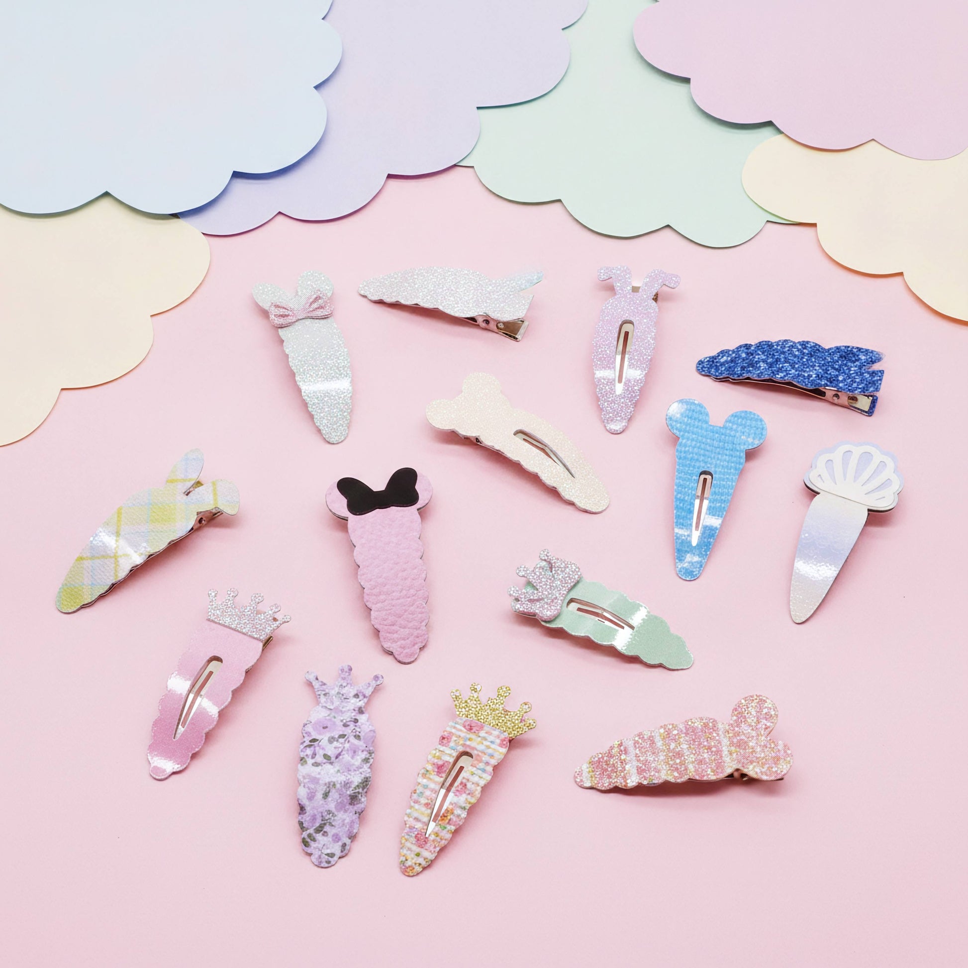 A set of pastel-colored hair clips arranged on a pink background with decorative cloud-shaped paper cutouts. The clips feature various designs including bows, flowers, shells, and glittery patterns in pink, blue, white, and yellow.