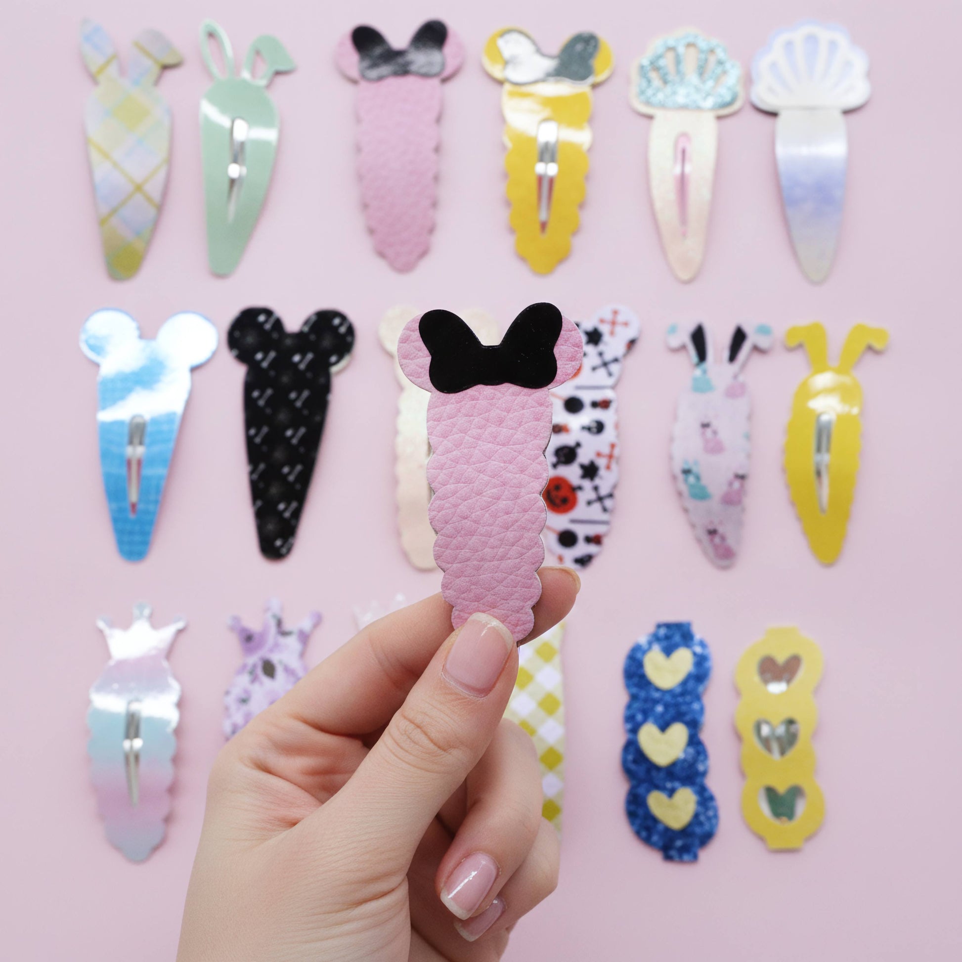 A hand holds a pink textured hair clip with a black bow, surrounded by other colorful hair clips in various designs against a light pink background.