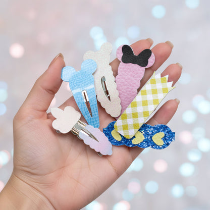 A hand holding five decorative hair clips shaped like cartoon characters and animals, including Minnie Mouse, Daisy Duck, and other playful designs, against a soft, blurred bokeh background.