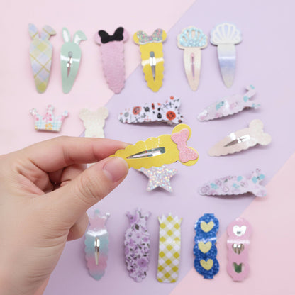 A hand holds a pastel yellow hair clip with a pink bow, shown among other pastel decorative hair clips arranged on a pink and lavender background.