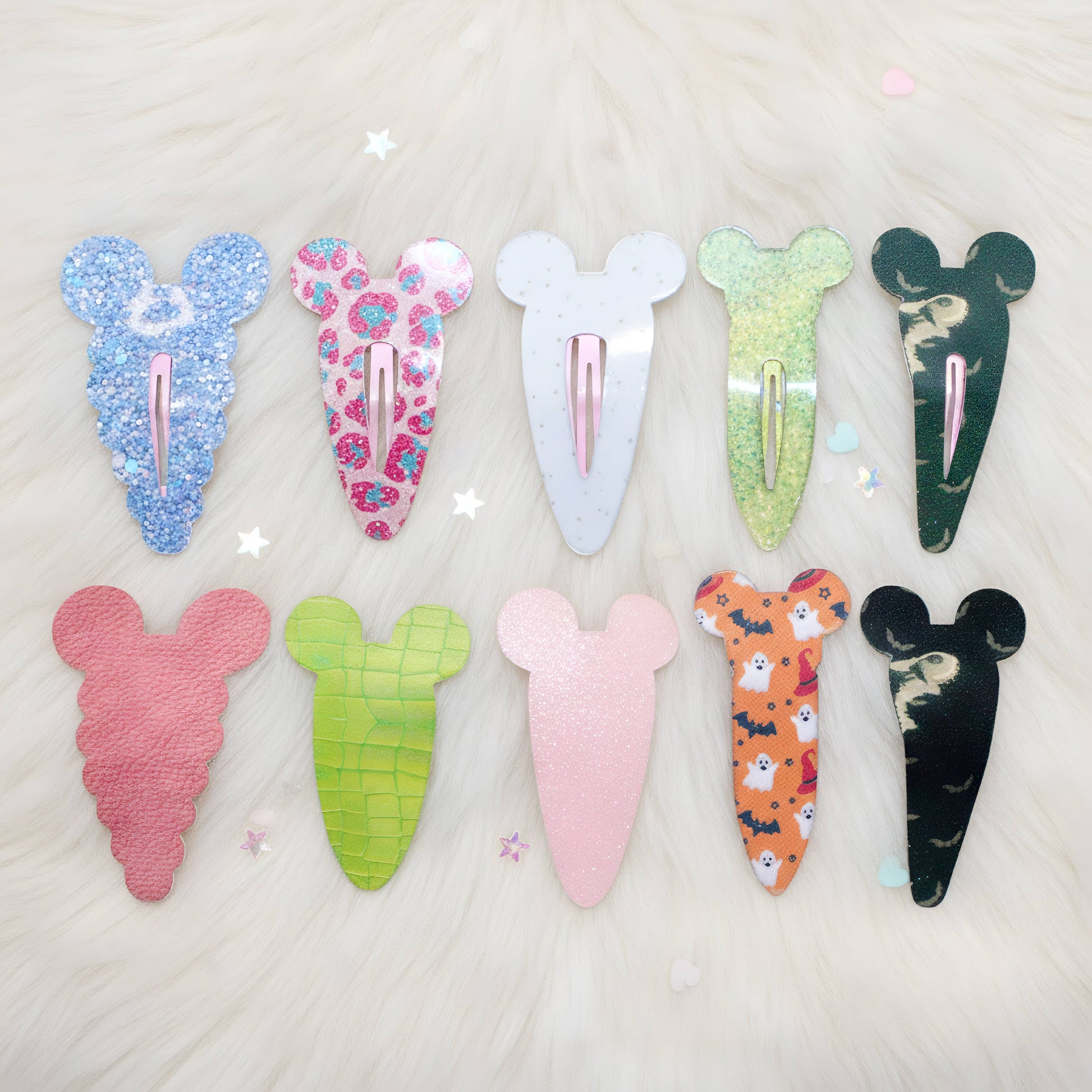 Eight decorative hair clips shaped like cartoon mouse ears, arranged in two rows on a white faux-fur background. The clips come in multiple colors and patterns, including glittery blue, floral pink, solid white, lime green, dark green, pink, green checkered, solid light pink, orange with cartoon prints, and black with a cat pattern.