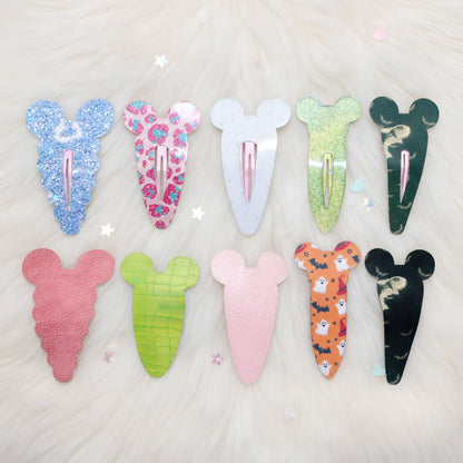 Eight decorative hair clips shaped like cartoon mouse ears, arranged in two rows on a white faux-fur background. The clips come in multiple colors and patterns, including glittery blue, floral pink, solid white, lime green, dark green, pink, green checkered, solid light pink, orange with cartoon prints, and black with a cat pattern.