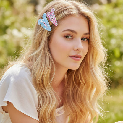 A blonde woman wearing two decorative cartoon mouse earshair clips in pastel blue and pink, photographed outdoors with a soft blurred green background.