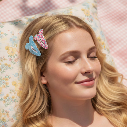 A close-up lifestyle shot of a young woman with blonde hair wearing two decorative  cartoon mouse hair clips, resting on a floral pillow with a pink gingham blanket in the background.