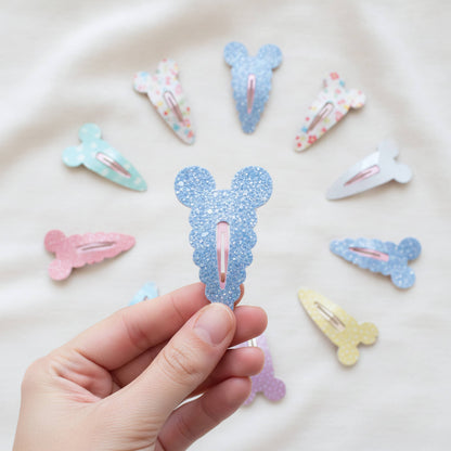 A hand holds a light blue glitter Mouse-shaped hair clip in the center, with several other pastel glitter and patterned hair clips arranged around it on a soft white fabric background.