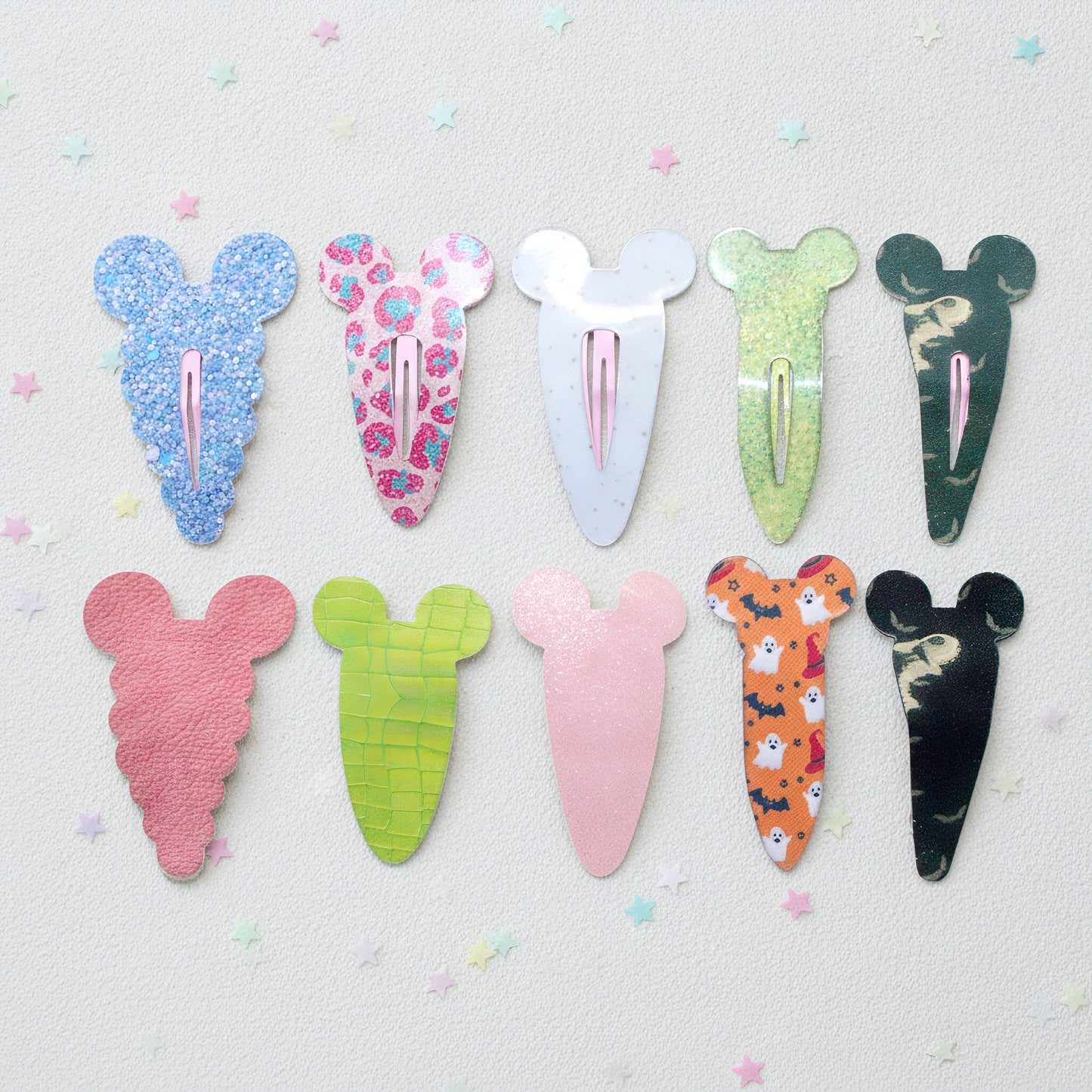 Eight hair clips shaped like cartoon mouse ears are arranged in two rows on a light textured background with small pastel star confetti. The clips feature various colorful patterns including glitter, floral, marble, and animal prints in shades of blue, pink, green, orange, and black.