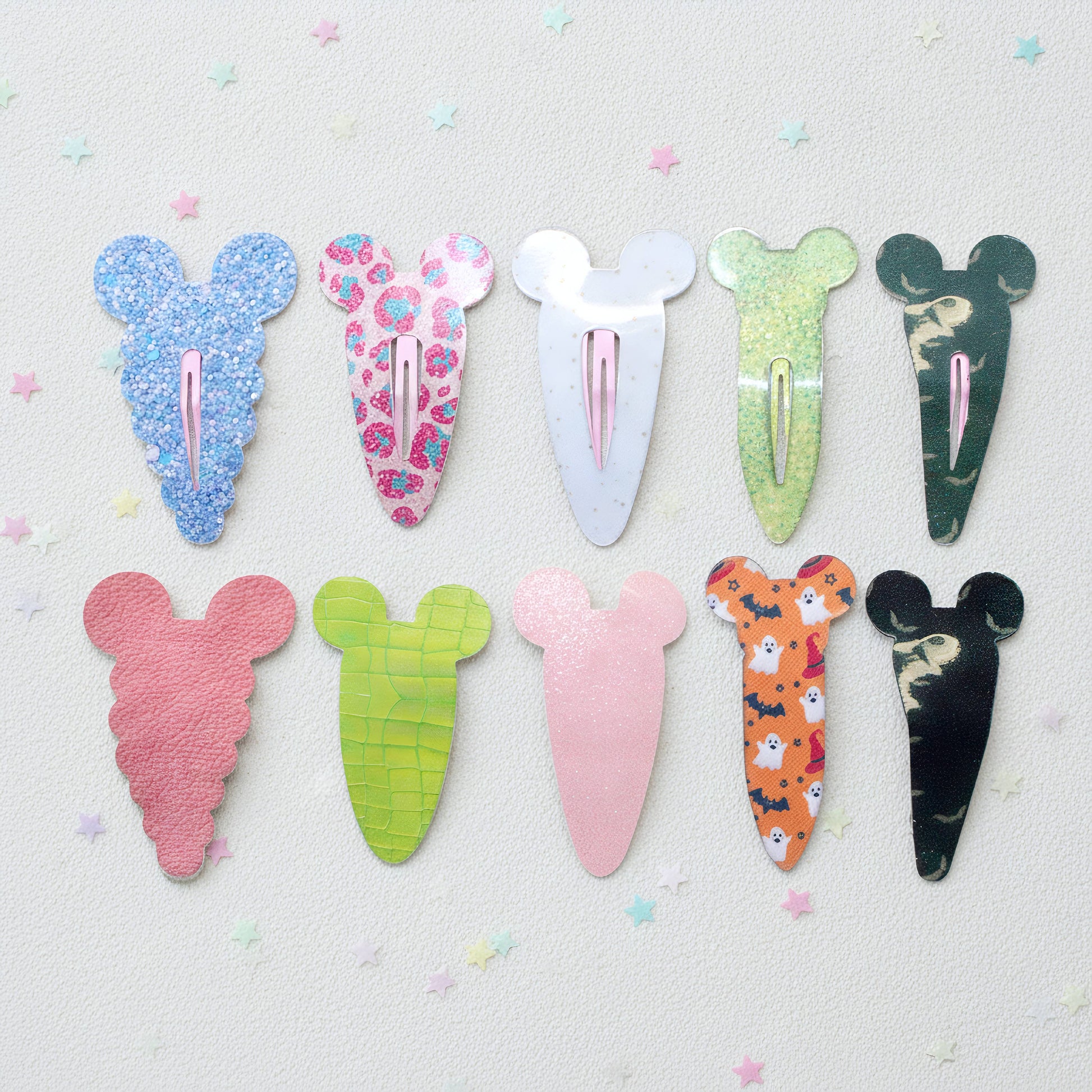 Eight hair clips shaped like cartoon mouse ears are arranged in two rows on a light textured background with small pastel star confetti. The clips feature various colorful patterns including glitter, floral, marble, and animal prints in shades of blue, pink, green, orange, and black.