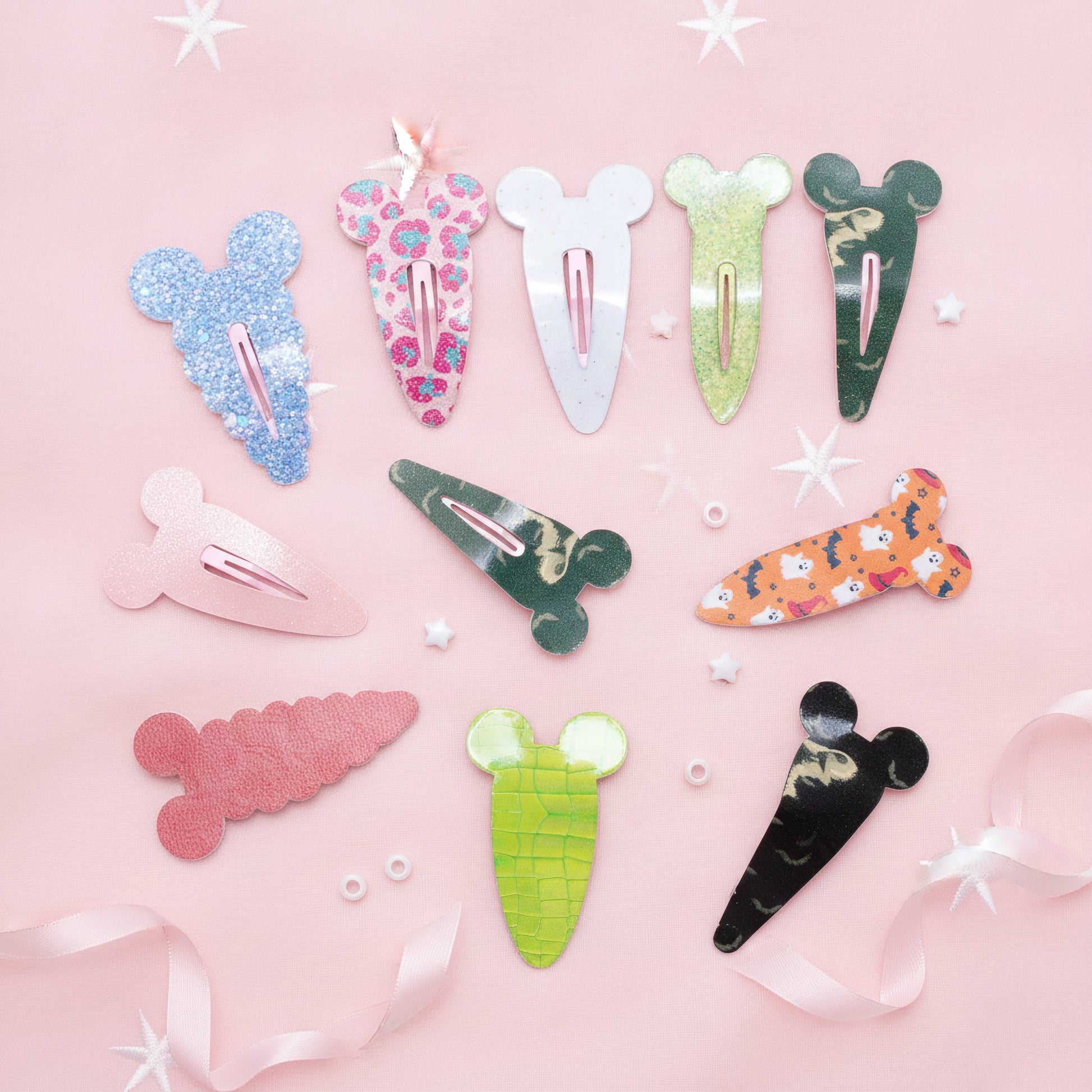A flat-lay of nine colorful hair clips shaped like cartoon characters and vegetables, arranged on a light pink background with white star stickers and pink ribbon accents.