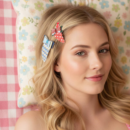 A blonde woman wearing three decorative hair clips, including a red strawberry clip, a blue bow clip, and a blue patterned clip, resting against a floral pillow and pink gingham blanket.