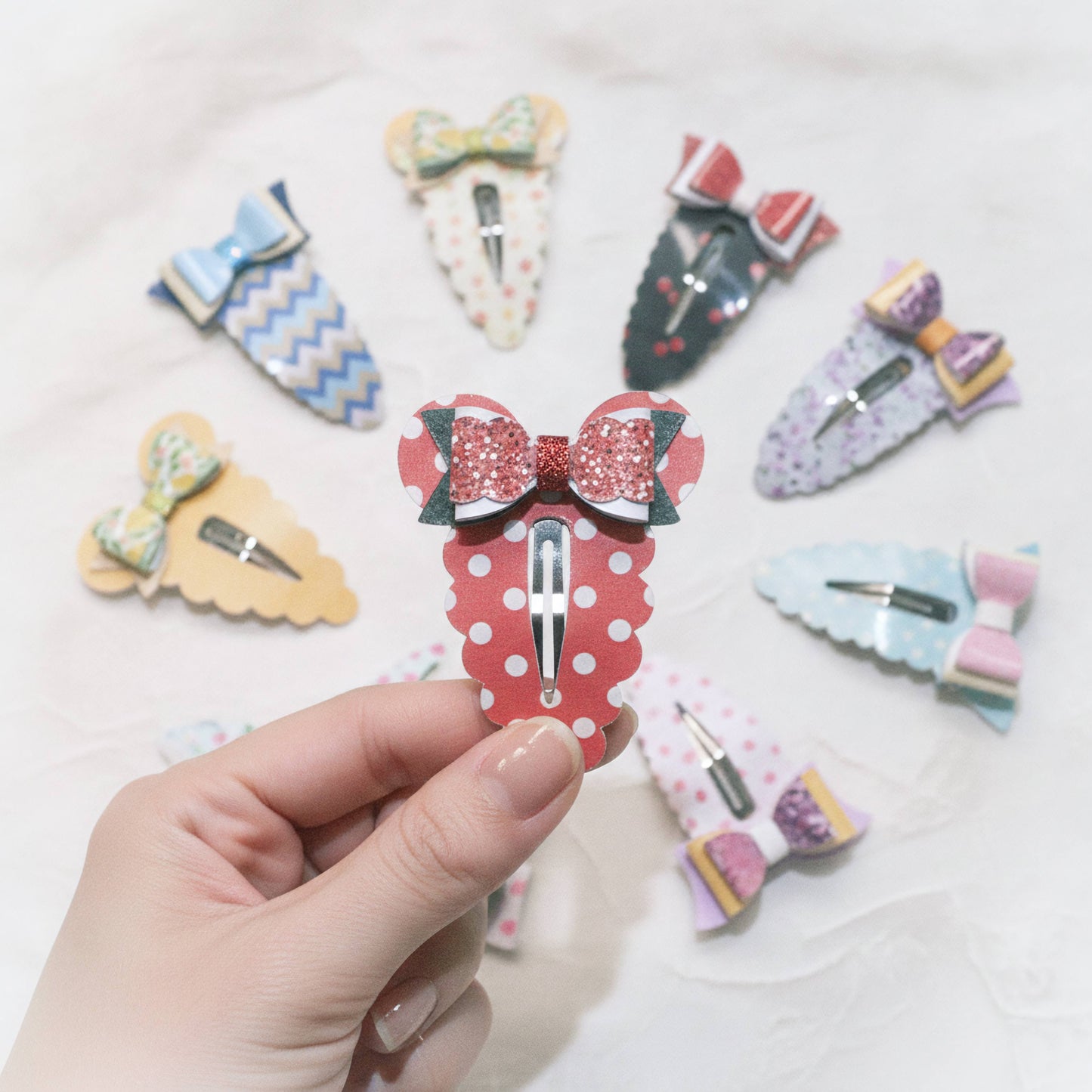 A hand holds a red and white polka dot hair clip with a glittery red bow, surrounded by other decorative bow hair clips on a light textured background.