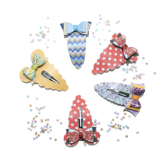 Five decorative hair clips arranged on a white background with scattered pastel star confetti. The clips include pastel blue-and-white chevron, red-and-white polka dot, green-and-white floral, lavender-and-white floral, and red glitter bow designs, each with a silver metal clip fastener.