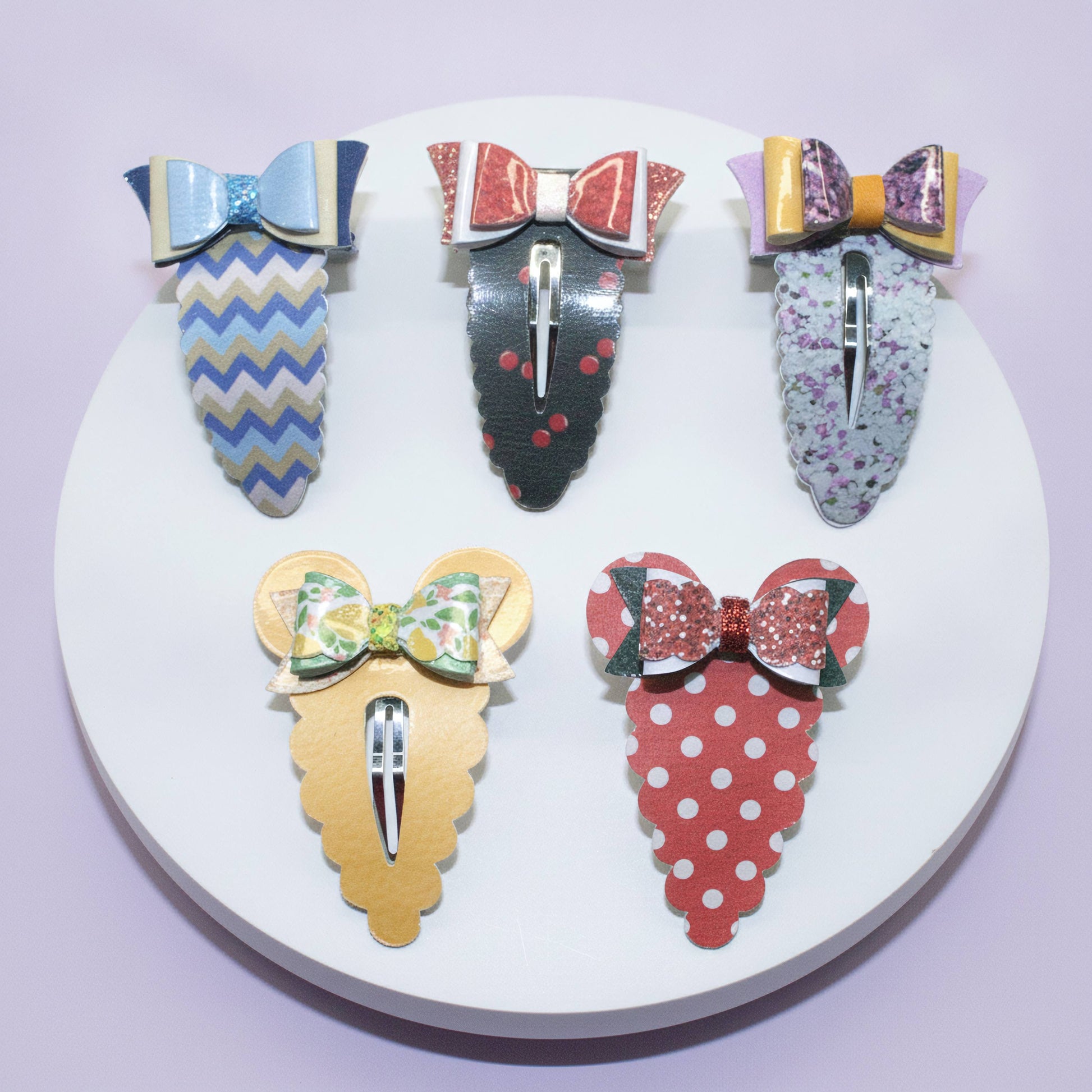 Five decorative hair claw clips arranged on a round white display stand against a pale lavender background. The clips feature colorful patterns including blue and beige chevrons, red and black polka dots, floral print, purple glitter, green and yellow floral, red and white polka dots, and a gold mouse-ear silhouette with a green floral bow.
