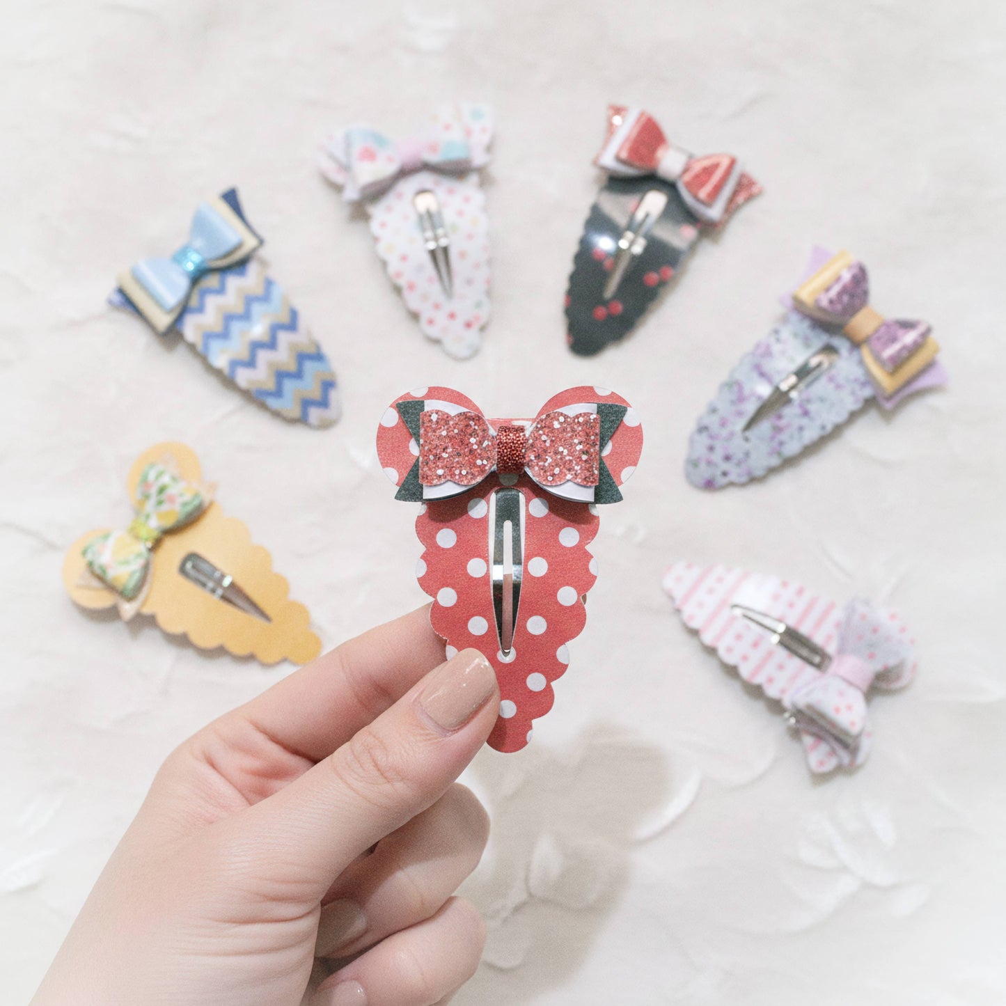 A hand holds a red and white polka dot hair clip with a glittery red bow, surrounded by six other decorative hair clips in various patterns on a white textured background.