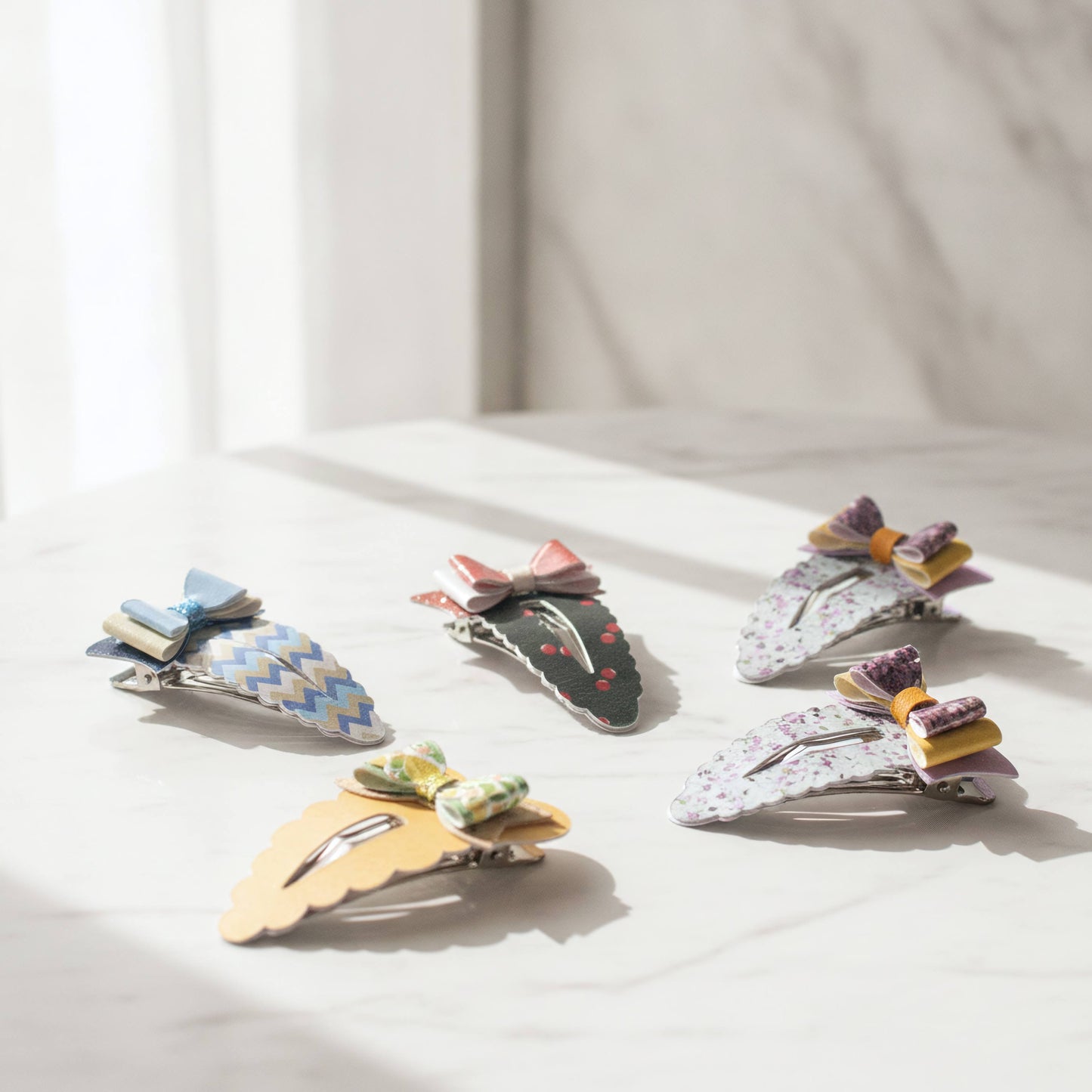 Five decorative hair claw clips arranged on a light marble surface with soft natural light. The clips feature colorful patterns and small ribbon bows, including blue-and-white chevron, dark green with red cherries, white floral, yellow scalloped, and pastel striped designs.