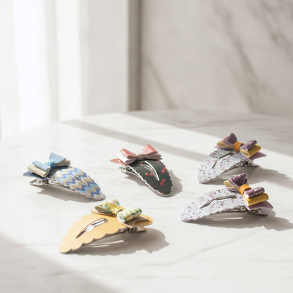 Five decorative hair claw clips arranged on a light marble surface with soft natural light. The clips feature colorful patterns and small ribbon bows, including blue-and-white chevron, dark green with red cherries, white floral, yellow scalloped, and pastel striped designs.
