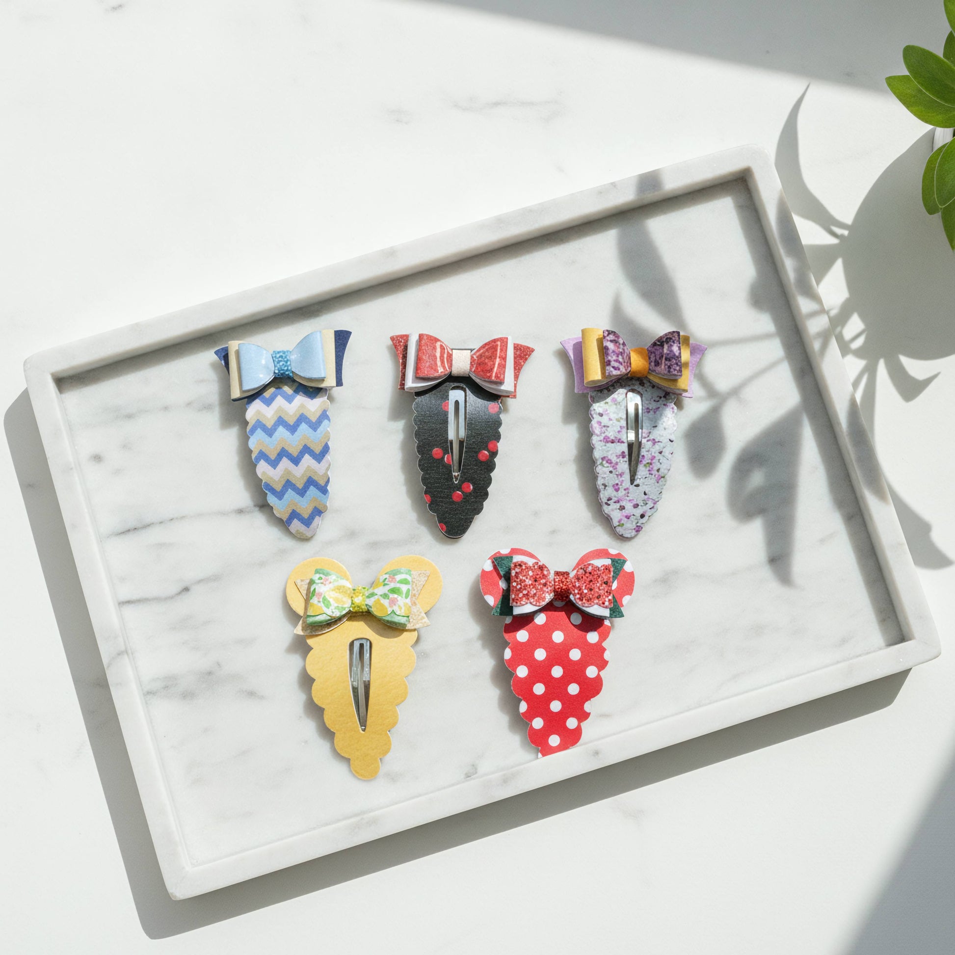 Five colorful hair claw clips arranged on a white marble tray, featuring patterned bows and scalloped bodies in blue, red, yellow, black, and white.