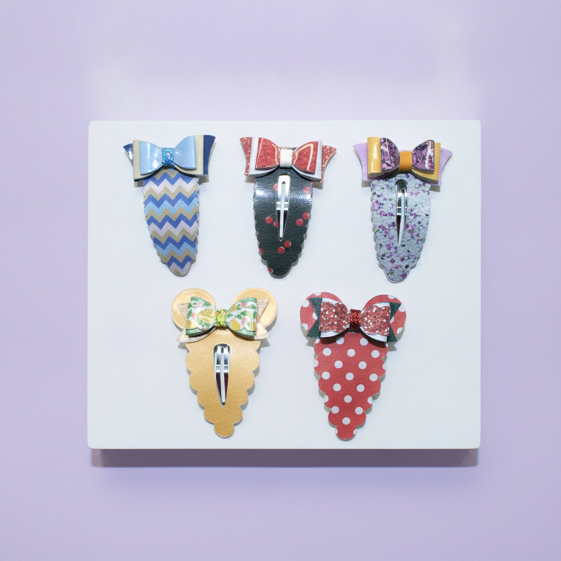 Five decorative hair claw clips arranged on a white rectangular display board against a light lavender background. The clips feature colorful patterned and bow designs in blue, black, purple, yellow, green, red, and white.