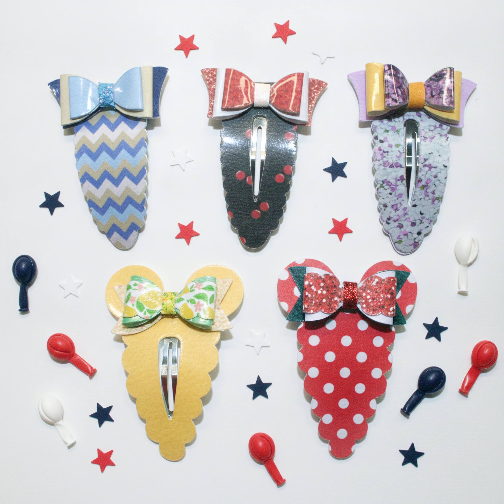 Five decorative hair claw clips arranged on a white background with scattered star and spoon-shaped props. The clips have colorful patterned designs: blue and beige chevron, red and pink bow, purple floral, yellow and green bow, and red-and-white polka dot with green bow accents.