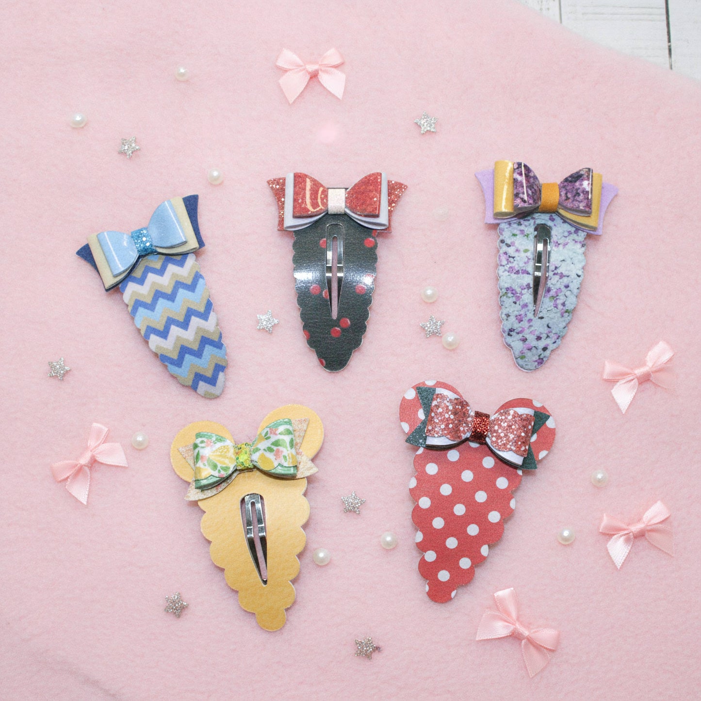 Five decorative hair claw clips arranged on a pink background with scattered pearl-like beads and small star confetti. The clips have colorful patterned bodies and bow details in blue, red, yellow, green, and purple.