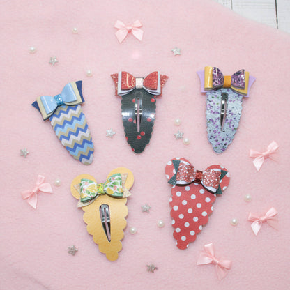 Five decorative hair claw clips arranged on a pink background with scattered pearl-like beads and small star confetti. The clips have colorful patterned bodies and bow details in blue, red, yellow, green, and purple.