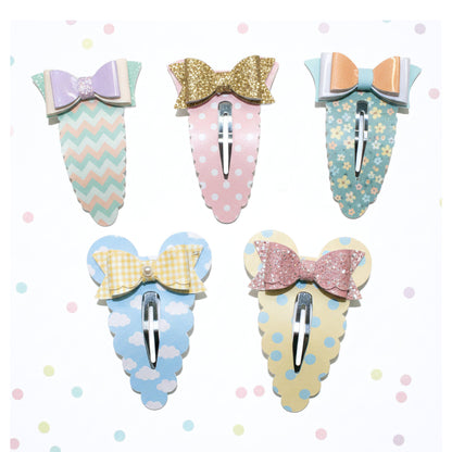 Five pastel hair claw clips arranged on a white background with scattered pastel dots. The clips feature bow and floral patterns in soft colors including mint, pink, peach, blue, yellow, and glittery gold.