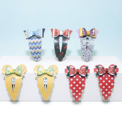 Six decorative hair claw clips displayed on a white stand against a light blue background. The clips feature colorful patterns and bows, including blue and white chevron, red and black polka dots, floral print, yellow and green patterned bow, red and white polka dots, and red strawberry print with green leaves.