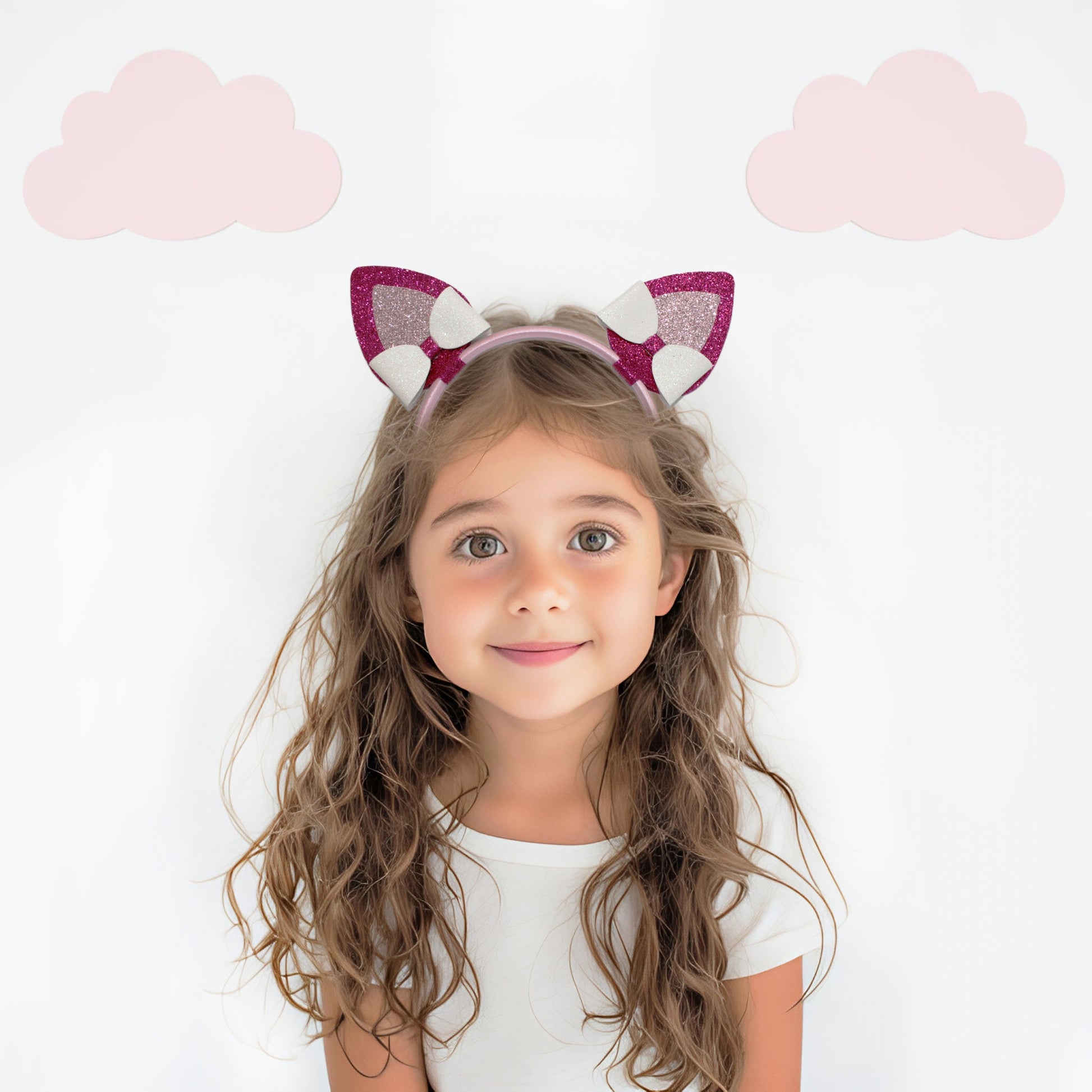 A cute young girl with wavy brown hair, wearing a white shirt and a pink cat ear headband with pom-poms, stands against a white background. Two pink cloud cutou