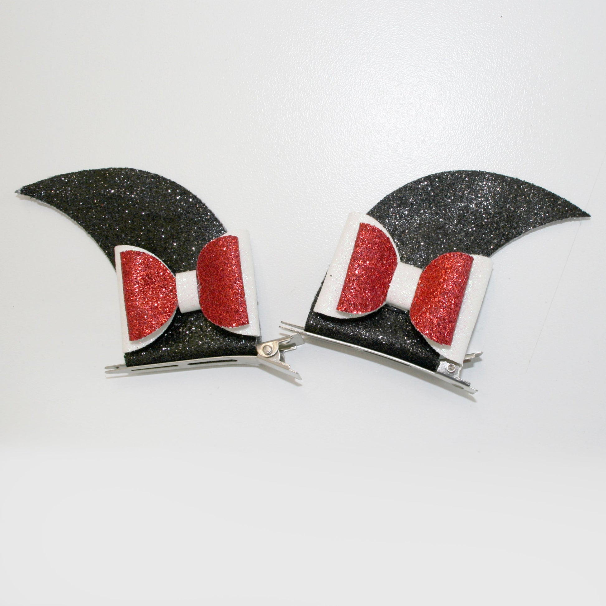 Photograph of two black hair clips with glittery red bows and white bases, positioned against a plain white background. The clips are designed in a sleek, modern style.