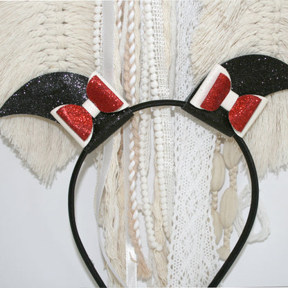 Photograph of a black headband with glittery red bows and bat-like ears, set against a plain white background. The headband has a smooth, matte texture, and the bows are sparkly and reflective.