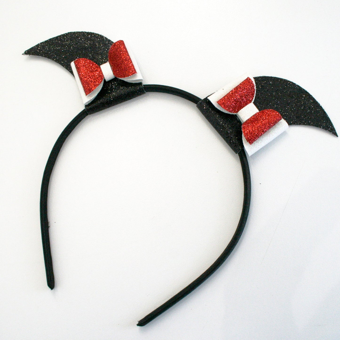 Photograph of a black headband with glittery red bows and bat-like ears, set against a plain white background. The headband has a smooth, matte texture, and the bows are sparkly and reflective.